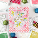 A handmade Christmas greeting card featuring a pink and white patterned background, a large layered pink bow, green and yellow mistletoe, and gold foil text reading 'Wishing you all the joys of another Christmas season.' The card is styled in a flat lay with craft supplies, glitter ornaments, and ink pads around it.