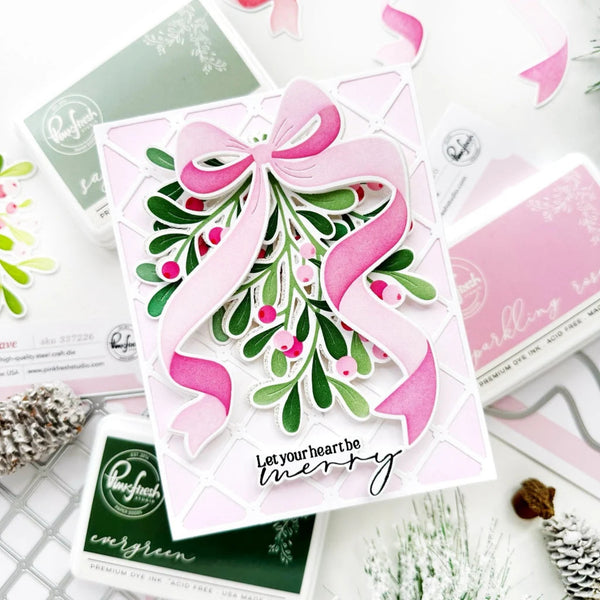 A handmade layered card featuring a large pink ribbon bow, green mistletoe with pink berries, and the text 'Let your heart be merry' on a white background, surrounded by Stampfing Ink products and holiday decorations.