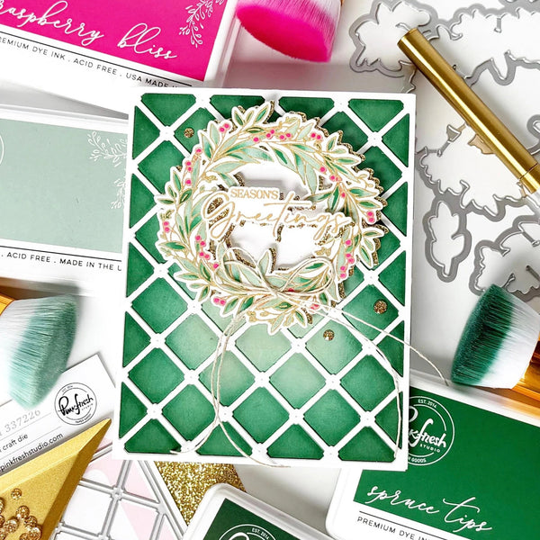 A handmade greeting card featuring a green and white lattice pattern with a die-cut wreath overlay that reads 'Seasons Greetings.' The card is styled in a flat-lay arrangement with crafting tools, brushes, and branded product packaging around it.