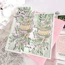 A handmade layered greeting card featuring two ornate Christmas ornaments, green leaves, and pink berries on a light green textured background, styled with white flowers and craft supplies.