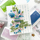 A handmade Christmas greeting card featuring a colorful stamped ornament, holly leaves, pinecones, and the phrase 'May my blossom this Christmas' in green script, styled on a white surface with crafting supplies.