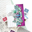 A handmade greeting card with a layered Christmas holly and ornament design, styled on a white surface with stamp blocks, ink pads, and patterned paper around it.