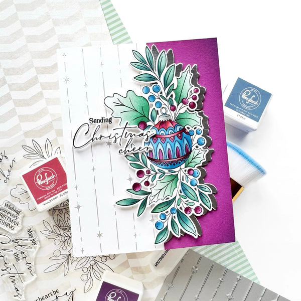 A handmade greeting card with a layered Christmas holly and ornament design, styled on a white surface with stamp blocks, ink pads, and patterned paper around it.