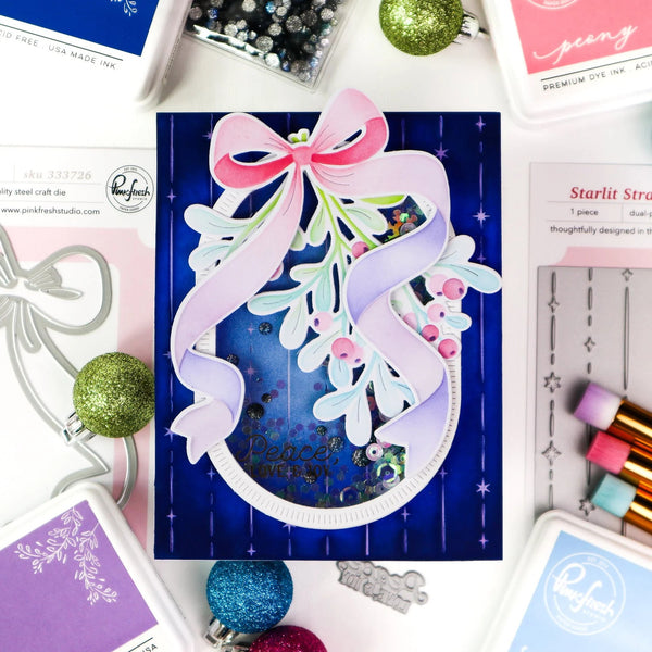 A handmade layered card featuring a dark blue background with vertical stripes, a large pink and lavender ribbon bow, white leafy branches, and small pink berries. The card is styled in a flat-lay with craft supplies, stamps, and ink pads around it.
