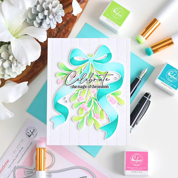 A handmade greeting card featuring a layered paper bow and mistletoe design with the text 'Celebrate the magic of the season,' styled in a flat lay with craft supplies, flowers, and teal paper on a white background.