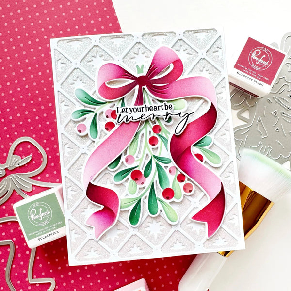 A handmade Christmas greeting card featuring a large pink ribbon bow, green holly leaves, red berries, and the phrase 'Let your heart be merry' in black script. The card is styled on a white surface with pink polka-dot paper, metal die-cut shapes, and Ranger ink pads visible around it.