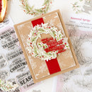 A handmade layered Christmas greeting card with a kraft paper base, a red ribbon, and a die-cut wreath with red berries and a red banner reading 'May your heart be filled with Christmas joy.' The card is styled on a white surface with clear stamps, floral stamp sheets, and a small gold dish visible around it.