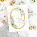 A handmade Christmas greeting card with a white background, gold glitter oval border, green and red berry foliage, and the phrase 'MAY YOUR heart BE FILLED WITH Christmas, JOY'. The card is styled in a flat-lay arrangement with gold and white glitter stars, a glass jar, and a card blank sheet in the background.