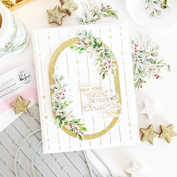 A handmade Christmas greeting card with a white background, gold glitter oval border, green and red berry foliage, and the phrase 'MAY YOUR heart BE FILLED WITH Christmas, JOY'. The card is styled in a flat-lay arrangement with gold and white glitter stars, a glass jar, and a card blank sheet in the background.