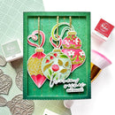 A handmade Christmas ornament card set displayed in a green presentation box, featuring three colorful ornament cutouts with floral and leaf patterns, gold hanging strings, and a die-cut sentiment reading 'Warm wishes this Christmas.' Surrounding props include stamp pads, embossed metal ornaments, and a makeup brush on a white background.