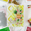 A handmade Christmas greeting card featuring a die-cut ornament, holly leaves, red berries, and the words 'May joy come this Christmas,' styled on a white surface with crafting tools and stamps around it.