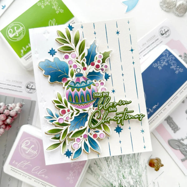 A handmade Christmas greeting card featuring a colorful stamped ornament, holly leaves, pinecones, and the phrase 'May my blossom this Christmas' in green script, styled on a white surface with crafting supplies.