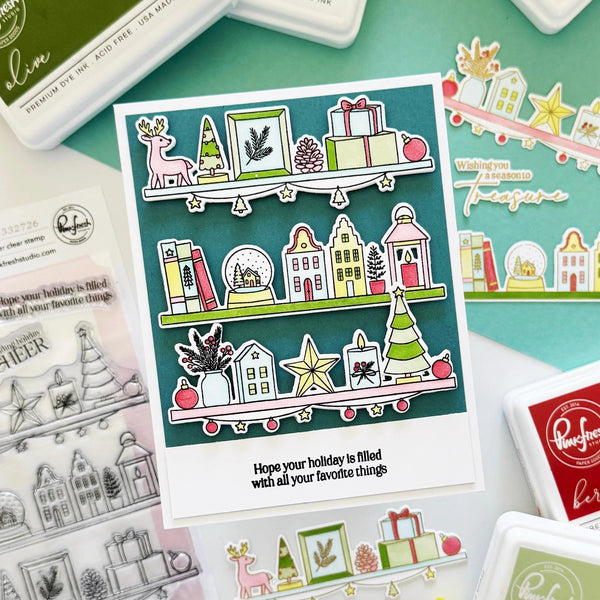 A handmade holiday-themed sticker card displayed on a mint green background with various crafting tools and stamps. The card features three illustrated shelves with Christmas decorations, houses, gifts, and stars, along with the stamped message 'Hope your holiday is filled with all your favorite things'.