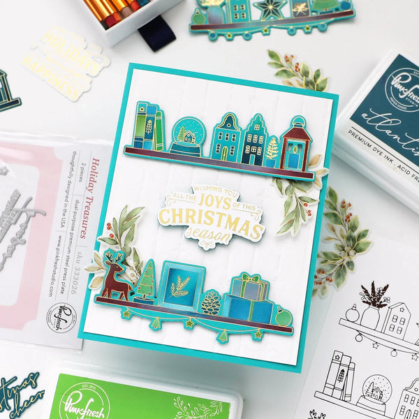 A handmade layered card featuring a teal and white holiday cityscape with houses, a snow globe, a reindeer, pine trees, and gift boxes, layered over a white cardstock base. The card is styled on a flat-lay desk with craft supplies, including an ink pad, a stamp sheet, and other holiday-themed items.