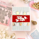 A handmade holiday card displayed on a light pink surface with crafting props around it. The card has a pale blue quilted background, a wide red center band with white layered die-cut text reading 'Wishing you holiday magic' and a bird, plus small white and gold Christmas motifs including reindeer, gift, snowflake, stocking, and ornaments.