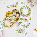 Watercolor-style botanical wreath stickers displayed on a white sheet and scattered around a shiny gold heart-shaped dish on a light pink surface.