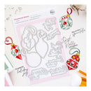 A flat-lay of a crafting kit featuring a clear stamp sheet with silver metal dies, surrounded by decorated Christmas ornaments and holiday-themed stamp designs on a white background.