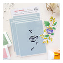 A flat lay of layered stamping mats with a black floral die-cut stamp design placed on top, alongside a branded card reading 'Festive Flourish' and a colorful stamped floral sample. The scene is styled on a light surface with small decorative props.