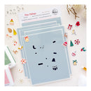 A set of layered acrylic stencils from Tiny Tidings, featuring Christmas-themed shapes like bells, gifts, pine trees, and snowflakes, displayed on a light beige background with craft tools and stamps.
