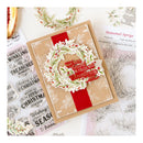 A handmade Christmas greeting card with a kraft paper base, a red ribbon, and a layered white and red wreath with holiday phrases, styled with stamp sheets and botanical stamps on a white background.