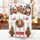 A festive Christmas greeting card displayed on a white wooden surface with a blurred holiday background. The card features a snowy house scene at the top, a white picket fence with red poinsettias, green wreaths with gold bows, two red birds, and potted Christmas trees with red flowers.