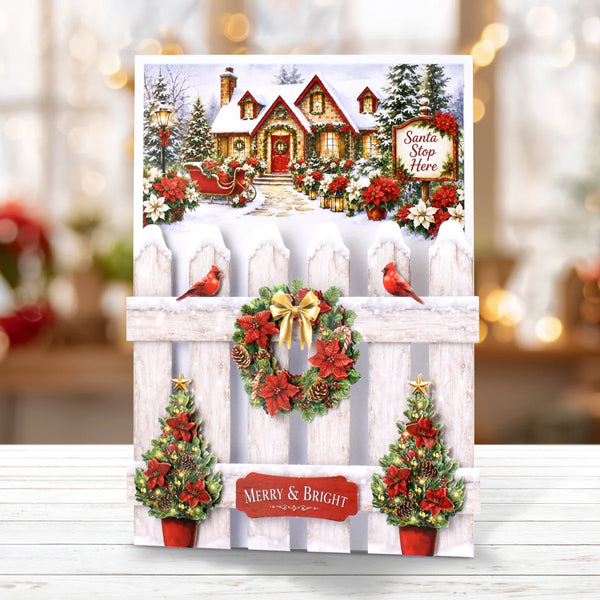 A festive Christmas greeting card displayed on a white wooden surface with a blurred holiday background. The card features a snowy house scene at the top, a white picket fence with red poinsettias, green wreaths with gold bows, two red birds, and potted Christmas trees with red flowers.