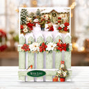 A festive Christmas greeting card displayed on a white wooden surface. The card features a snowy winter scene with a snowman, children, a decorated cabin, poinsettias, and a light green picket fence.