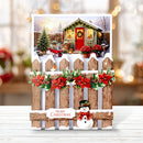 A festive Christmas greeting card or decorative sign featuring a snowy winter cabin scene printed on the top half, layered over a rustic wooden picket fence with red poinsettias, green leaves, pinecones, and a small snowman at the bottom. The sign reads “Merry Christmas” and is displayed on a light wood surface with a softly blurred holiday background.
