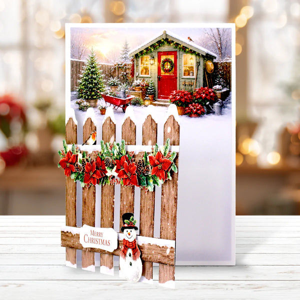 A Christmas greeting card featuring a snowy scene with a small cabin, decorated trees, red poinsettias, and a wooden picket fence with a snowman and 'Merry Christmas' banner, displayed on a white surface with a blurred festive background.