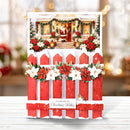 A festive Christmas greeting card featuring a snowy red picket fence with white poinsettias and green garlands, topped with a photo of Santa Claus at a decorated holiday porch. The card is displayed upright on a white wooden surface with a soft, blurred holiday background.