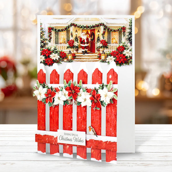 A Christmas greeting card featuring a red picket fence with white flowers and green garlands, with a printed holiday house scene above. The card is displayed standing upright on a white surface with a softly blurred festive background.