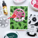 A handmade Christmas greeting card featuring a red cardinal and layered pink flowers on a green holly-patterned background, surrounded by crafting supplies including ink pads, a small bottle of dye, metal dies, and clear stamps.