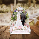 A wedding-themed tabletop display featuring a printed arch-shaped backdrop with a bride and groom seen from behind, surrounded by pink and white flowers, with a sign reading 'HAPPILY EVER AFTER,' set on a wooden surface against a softly blurred outdoor wedding background.