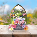 A 3D pop-up cardboard birthday card shaped like a garden arch, featuring a colorful floral landscape, a bluebird, and a base of blooming flowers. The card is displayed on a wooden surface with a blurred garden background.