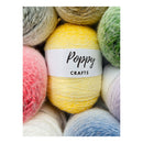 A close-up of several fuzzy yarn balls arranged together, with a central yellow ball wrapped in a white label that reads 'Poppy CRAFTS'. Other visible yarn colors include pink, green, blue, white, and gray.