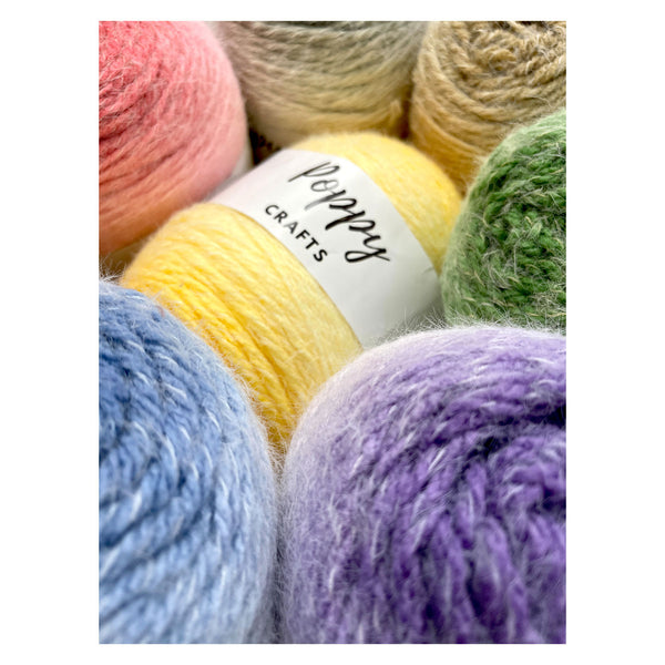 A close-up of several skeins of yarn in pastel colors, including pink, yellow, blue, purple, green, and beige, with a white label reading 'Poppy CRAFTS' wrapped around one yellow skein.