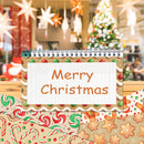 A spiral-bound notepad with a gingerbread border and the words 'Merry Christmas' on the cover, placed on top of holiday-themed patterned paper with a blurred Christmas tree and hanging ornaments in the background.