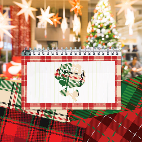 A spiral-bound notepad with a red plaid border and a white center panel featuring a green and red rose illustration, styled on top of red plaid fabric with a blurred Christmas tree and hanging star decorations in the background.