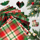 A styled flat-lay of Christmas-themed plaid paper napkins or paper products in red, green, black, and cream tartan patterns, arranged on a light gray surface with pine branches, ornaments, and small warm lights.