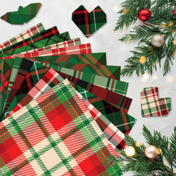 A styled flat-lay of Christmas-themed plaid paper napkins or paper products in red, green, black, and cream tartan patterns, arranged on a light gray surface with pine branches, ornaments, and small warm lights.