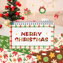 A festive Christmas-themed spiral notebook centered on a table, surrounded by holiday decorations including a decorated tree, ornaments, wrapped gifts, and patterned paper sheets. The notebook cover reads “MERRY CHRISTMAS” in red letters with a green and red plaid border.