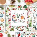 A styled flat-lay of Christmas-themed printed sheets or cards with a central illustration of two birds surrounded by holly, pine branches, candy canes, ornaments, and snowflakes, surrounded by holiday props like a coffee cup, keyboard, pinecone, and wrapped gift.