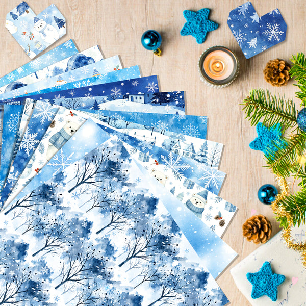 A styled flat-lay of blue winter-themed patterned paper sheets fanned out on a light wood surface, surrounded by holiday decorations including pine branches, blue star ornaments, a lit candle, and small gift boxes.