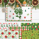 A flat-lay display of Christmas-themed paper products, including an open notebook with a green rose illustration and a green plaid border, alongside four sheets of patterned paper with festive designs like pinecones, poinsettias, and Christmas trees. The scene is decorated with pine branches, dried orange slices, and wooden star ornaments on a white wood background.