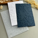 Three embossed floral cardstock sheets in white, navy blue, and a translucent clear sheet, arranged on a neutral gray surface with dried floral props.