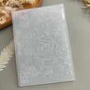 A translucent acrylic or plastic stamping plate with embossed floral and butterfly designs, displayed on a neutral gray surface with dried botanical props.