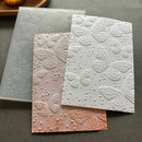 Three embossed butterfly cardstock sheets in white, pale pink, and peach, layered over a translucent silicone stamping plate on a gray surface.