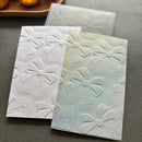 Two embossed paper sheets with a repeating bow pattern, one in white and one in pale green, displayed on a gray surface.