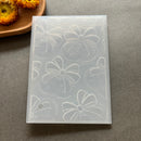A translucent silicone mold with embossed bow patterns, displayed on a gray textured surface with a wooden tray and dried flowers in the background.