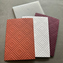 Three textured paper sheets with a raised geometric diamond pattern, shown in terracotta orange, white, and deep burgundy, with a translucent clear sheet behind them on a gray background.
